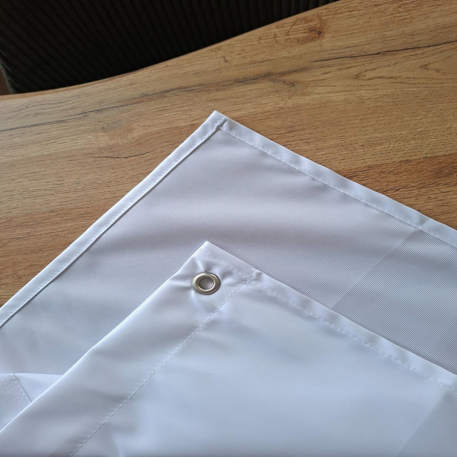 Ein einzelnes weißes Stoffbanner liegt auf einem Holztisch; in der Ecke ist eine silberne Metallöse deutlich sichtbar, und die sauber genähten, verstärkten Kanten heben sich durch ihre präzisen Nähte vom glatten, leicht glänzenden Material ab.